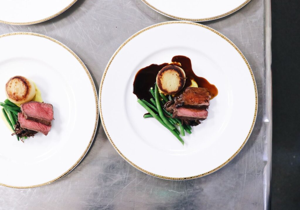 Two identical fine dining plates on a kitchen pass, each bearing sliced roasted beef, pan-fried polenta, green beans and a rich dark jus, ready to be served