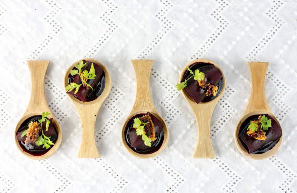 Row of wooden spoons each holding a single dark bite-sized amuse-bouche topped with micro-herbs, representing the chef's introductory course