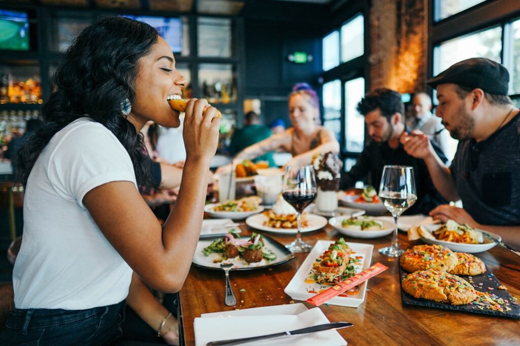 A group of friends eating and laughing together at a lively casual dining restaurant, sharing plates of food with wine glasses on a wooden table