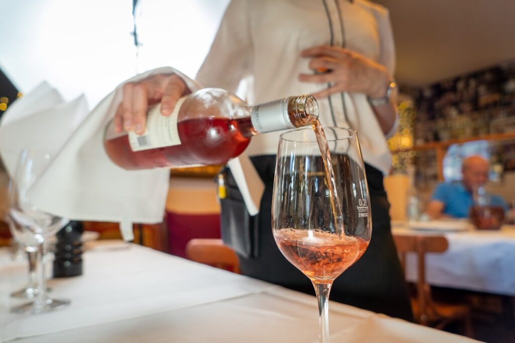 A server in a white apron pours rosé wine into a large crystal glass at a restaurant table