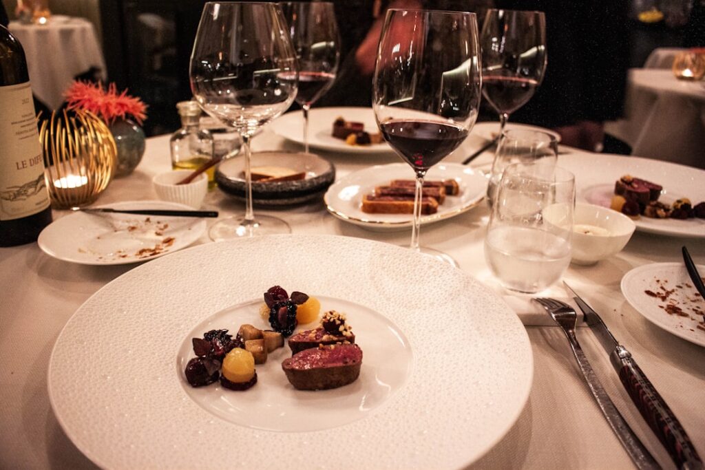 A precisely plated fine dining tasting menu course on a white plate, surrounded by red wine glasses and a candlelit table setting