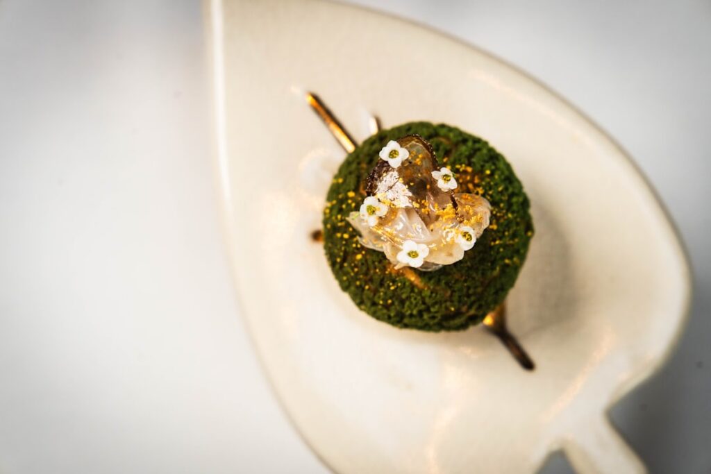 A single-bite amuse-bouche: a jewel-like morsel of herb-crusted food topped with edible white flowers and gold leaf on a leaf-shaped ceramic dish