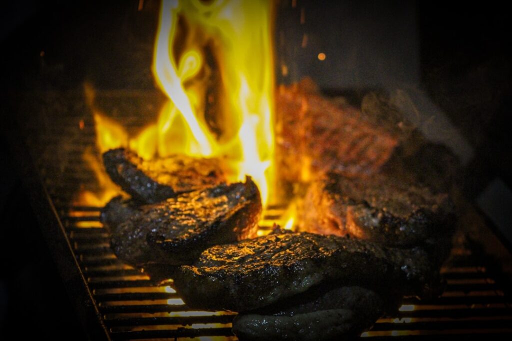 Steaks cooking over open flames on a grill with heavy char crust developing, matching the smoky notes of Northern Rhone Syrah