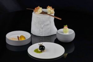 Three artistic tasting menu courses presented in sculptural vessels against a dark background, illustrating the theatrical progression of a fine dining tasting menu