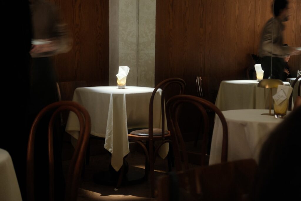 Dimly lit fine dining room with white-clothed tables and candlelight, servers moving quietly between tables