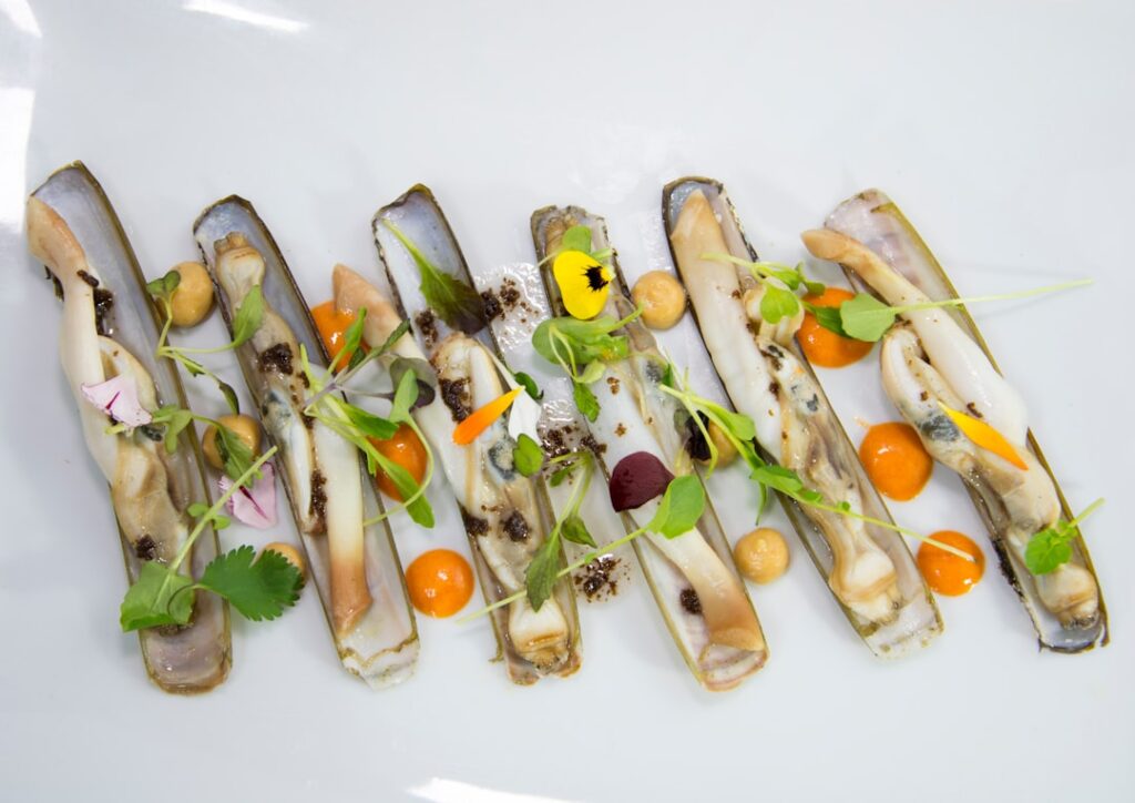 Elegantly plated razor clams with microgreens, cherry tomatoes, and edible flowers on a white plate